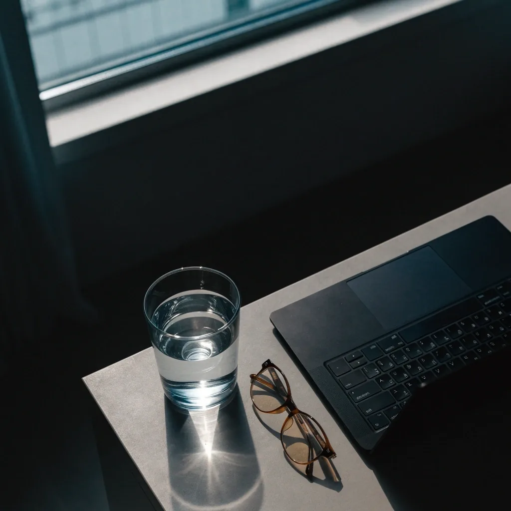 Matte counter with a closed laptop, a glass of water and reading glasses in cool daylight